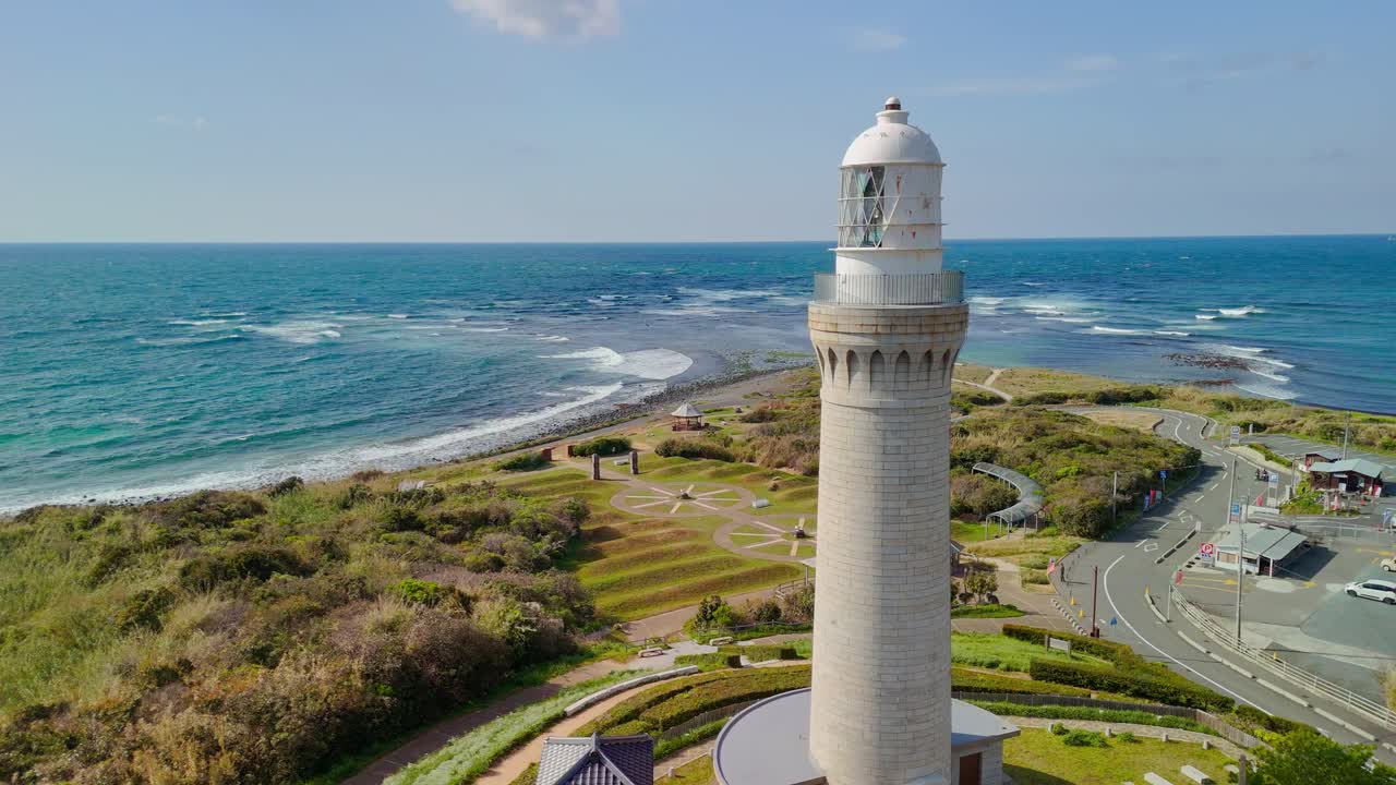 A beautiful, static aerial shot of a tall, white lighthouse overlooking the turquoise ocean on the scenic coast of Japan, with a park and visitor area below.