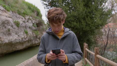 teenage boy texting on phone while walking along scenic river path