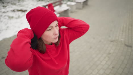 lady performing hand stretch by turning arm outdoors in winter fitness routine, dressed in athletic wear, snow-covered ground, frosted trees, benches in serene park setting