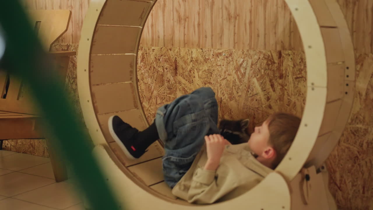young boy curled inside wooden wheel softly resting beside bench, black pet napping, warm plywood walls, hay backdrop, gentle quiet morning play scenes show child exploring cozy nook, curling into