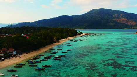 An-aerial-footage-of-long-and-white-sandy-beach-line,-with-unrecognisable-people-plus-boat-being-dock-or-park-by-the-beach-and-resort-int-the-background