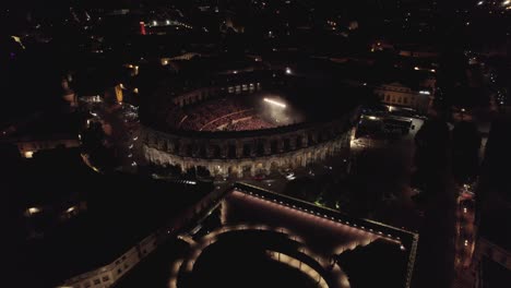 Drone-En-Las-Arenas-De-Nîmes-Y-El-Museo-Romano-En-Medio-De-La-Noche,-La-Gente-Está-Viendo-El-Concierto-Y-Hay-Luces-De-Varios-Colores