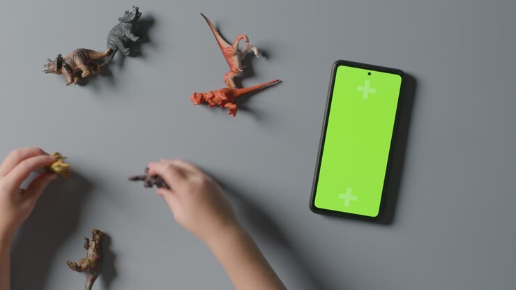 Ripresa dall'alto di un ragazzo che gioca con dinosauri giocattolo accanto a un telefono cellulare con schermo verde.