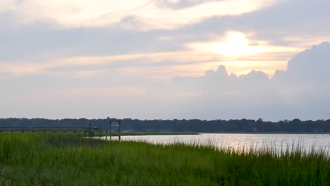 smooth dolly from left to right with parallax effect on foreground grass that reveals a coastal marsh at sunset with gorgeous colors in the clouds and a relaxing vibe