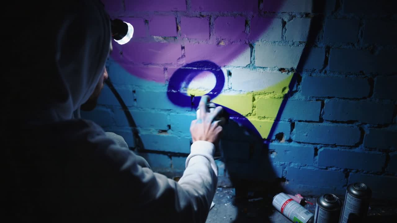 Lampebelysning belyser spraymalende kunstner i hættetrøje, der tegner løkker, der danner motiv på mursten, dåser, plads til tekst
