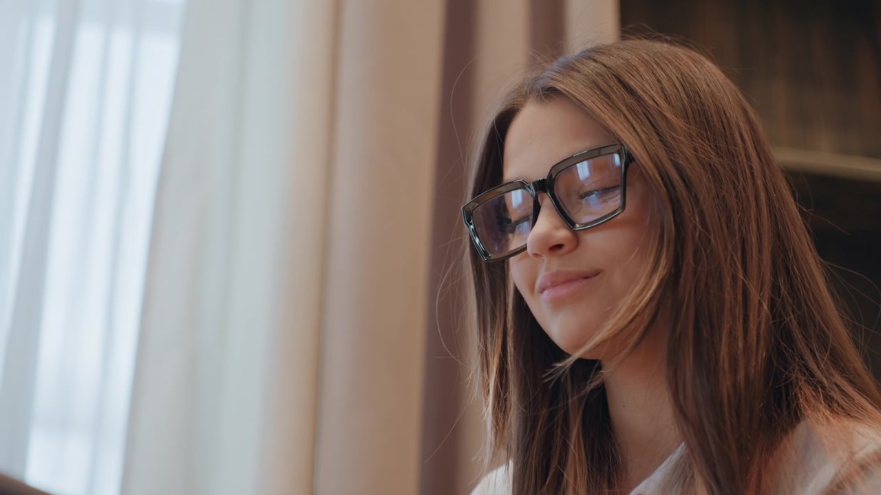Caucasian woman with glasses studying near window soft natural light, long brown hair, serious focused expression, typing on laptop, reading notes, freelance writer and researcher roles, quiet modern