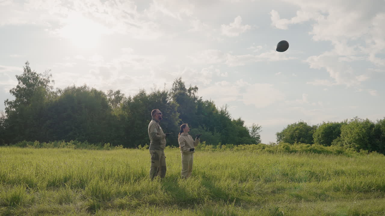 two field workers in protective uniform holding remote device while releasing tethered black balloon over expansive green meadow under partly cloudy sky during atmospheric survey exercise