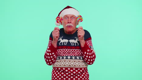 Grandfather-in-Christmas-sweater-holding-candy-striped-lollipops-hiding-behind-them,-fooling-around