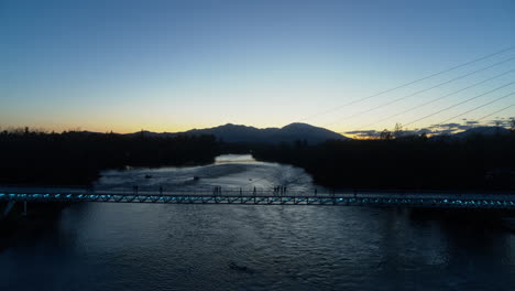 Aerial-pan-up-over-the-Sundial-Bridge-while-people-are-walking-across-it-in-the-dusk-of-sunset-over-the-Sacramento-River-in-Redding,-California