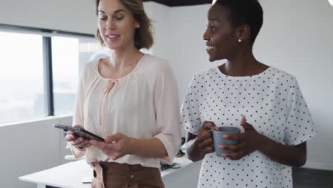 duas colegas mulheres diversas caminhando, olhando para o tablet e discutindo no escritório