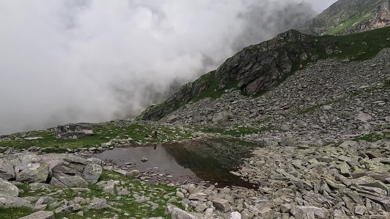 Un escursionista solitario accanto a un piccolo lago alpino, circondato da rocce, nebbia e ripidi pendii alpini nelle Alpi italiane.