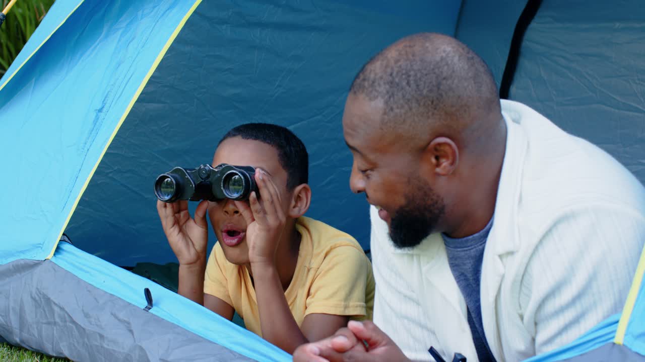 Father and boy responding to motion outside blue tent, boy in yellow shirt scanning with binoculars