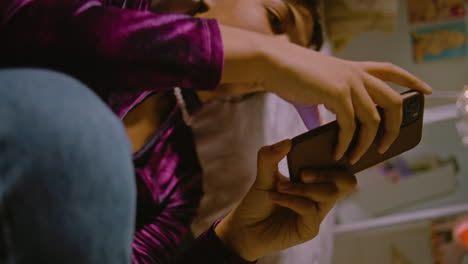 vertical view of african american girl chatting or scrolling social media networks using phone, sitting near the bed in her cozy bedroom. teenage girl spending leisure time and having fun at home.