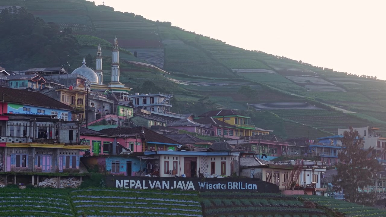 Wioska na zboczu góry otoczona gęstą zieloną roślinnością z napisem "Nepal Van Java" i malowniczym wiejskim krajobrazem uchwyconym w kinowym świetle dziennym. Nepal Van Java, Indonezja