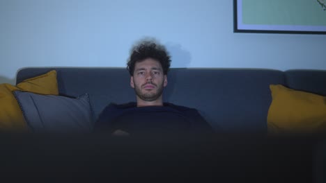 young tired male watching television at night while relaxing on couch