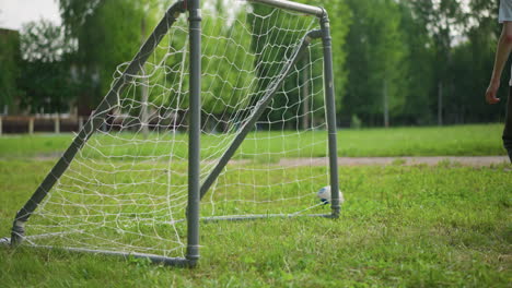 un niño pequeño patea con entusiasmo la pelota hacia un poste de la portería mientras su hermano mayor se mueve para interceptarla, en el fondo, la gente está borrosa