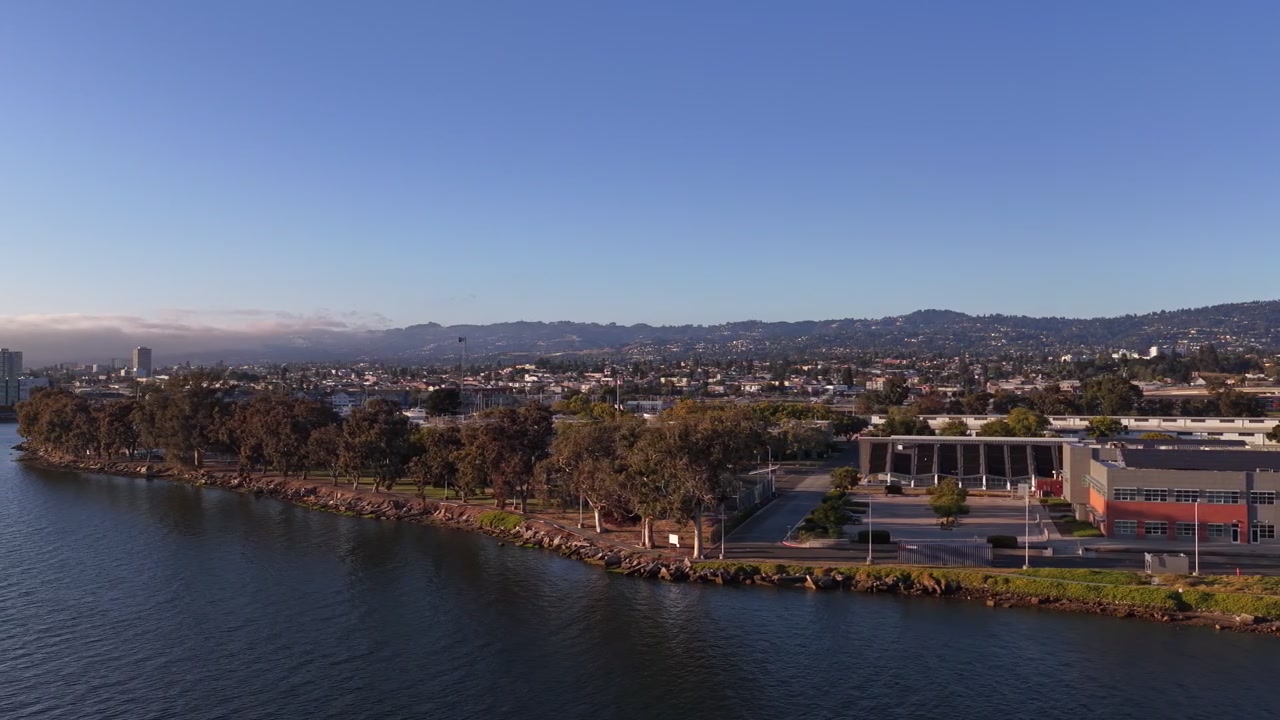 Vista aerea che sorvola il lungomare dell'Estuario di Oakland in California, mostrando un parco alberato, acqua blu calma e lo skyline della città sotto un cielo sereno e soleggiato al tramonto.