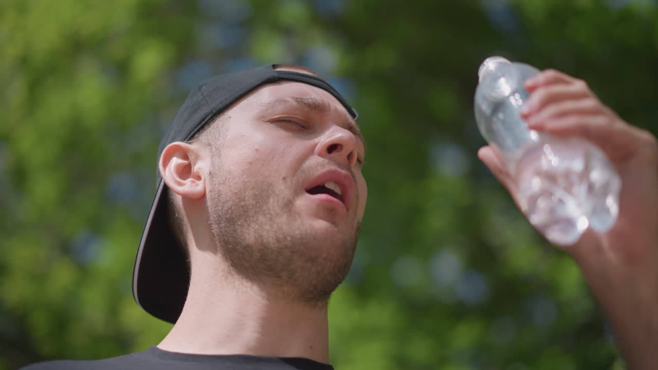 햇빛 아래에서 물을 마시는 남성, 밝은 빛 때문에 눈을 가늘게 뜨면서 플라스틱 병을 입으로 가져가는 남성의 중간 샷, 모자는 거꾸로 썼고, 잎이 무성한 공원 배경은 운동 후의 열기와 회복을 암시합니다.