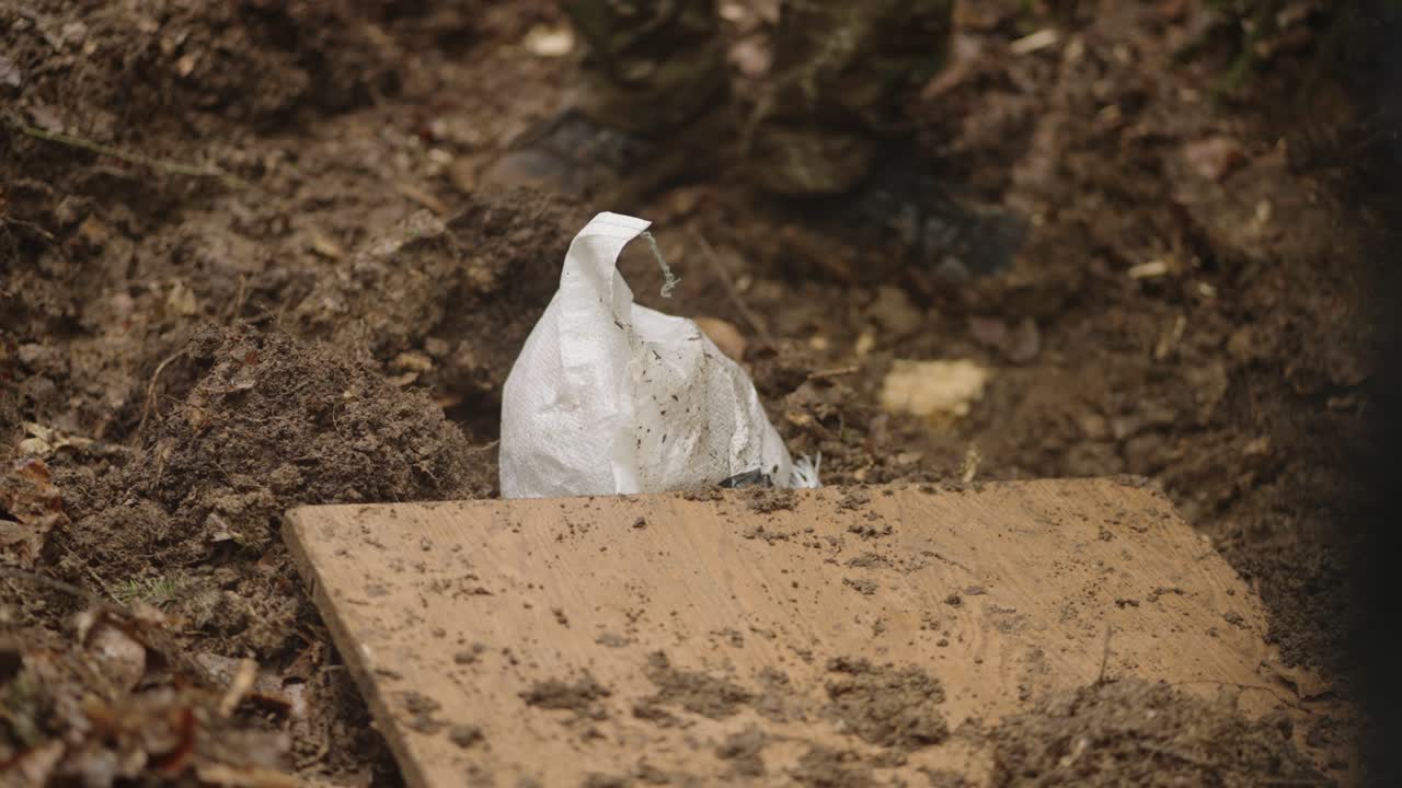 Close up of hooded captive man lying on dirt ground with hands tied as military personnel place wooden board over him