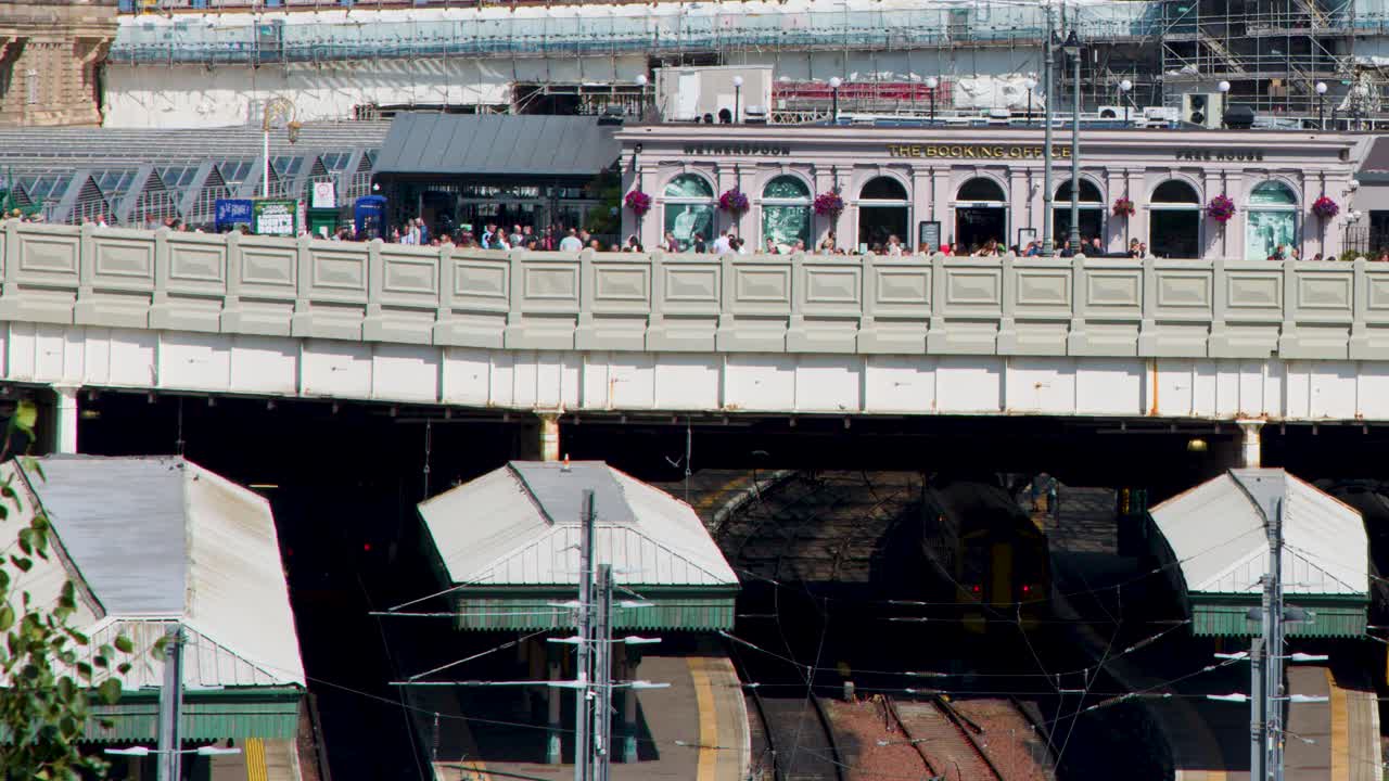 Yellow-fronted train enters busy urban railway station under bright daylight, static wide shot perspective