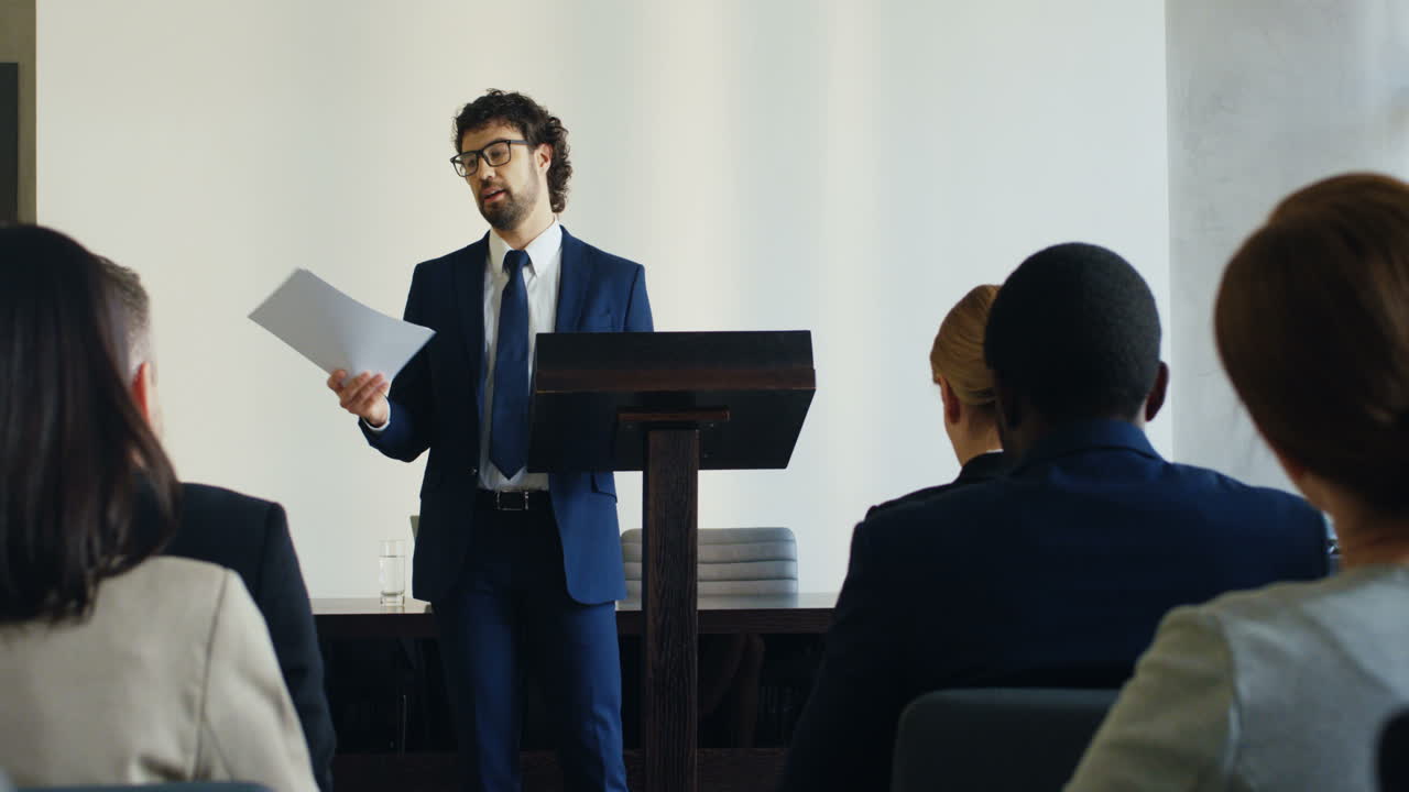 un hombre de negocios caucásico orador en un podio con ropa formal, sosteniendo documentos y hablando en una sala de conferencias frente a muchas personas