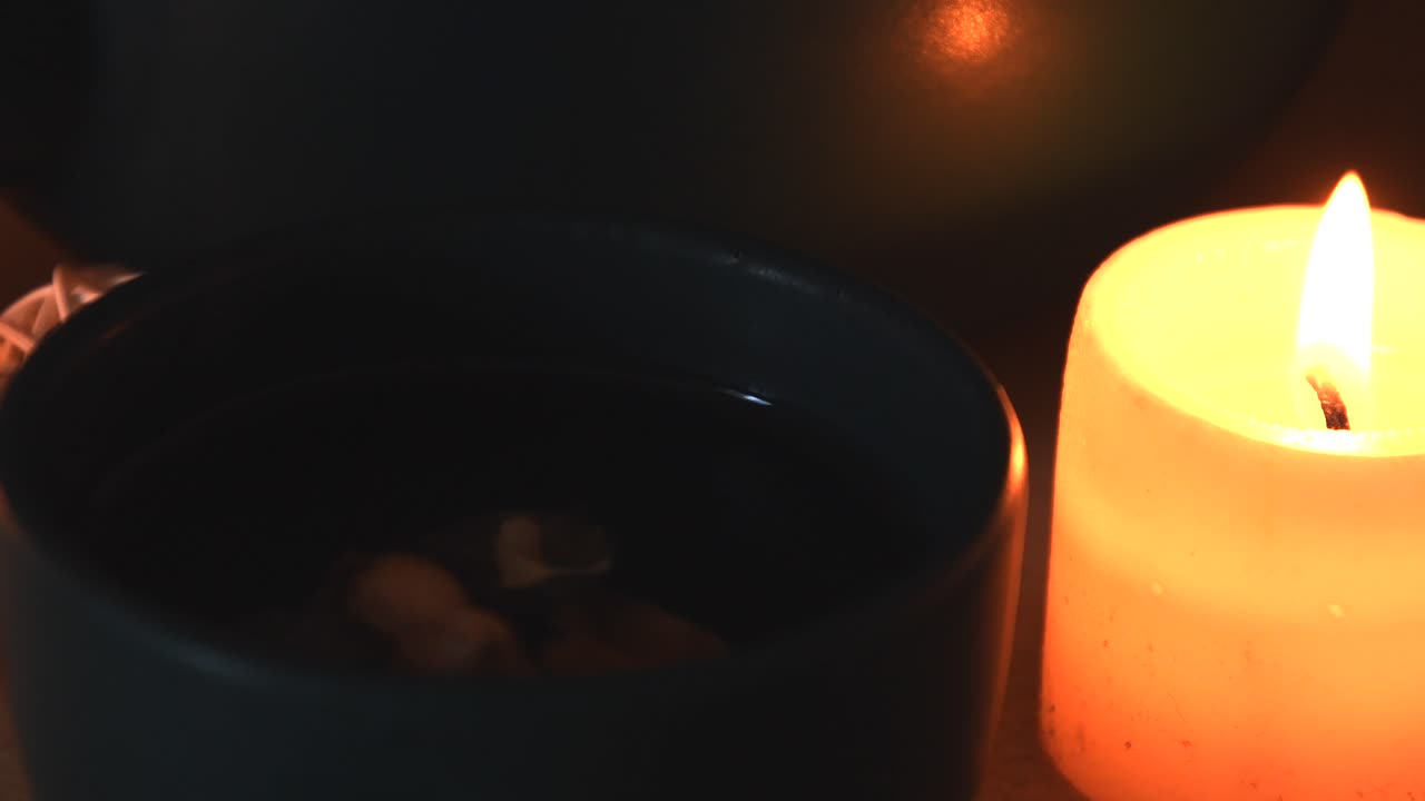 Avslappende detaljbilde i bakgrunnen av urtete i en grønn kopp, med damp som stiger opp, nær et stearinlys med en flakkende flamme, en tekanne og noen urter.