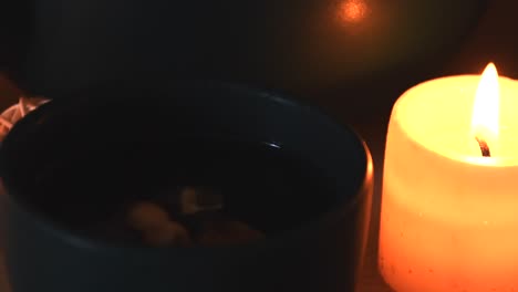 relaxing background detail shot of an herbal tea on a green cup, with steam coming out, near a candle with a flickering flame, a tea pot and some herbs