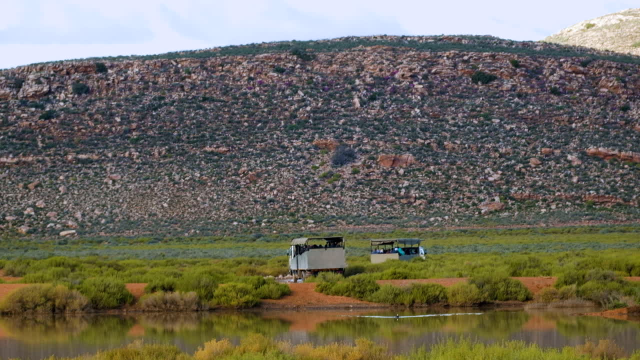 Safari trucks loaded with tourists heads out on a game drive, long shot over dam
