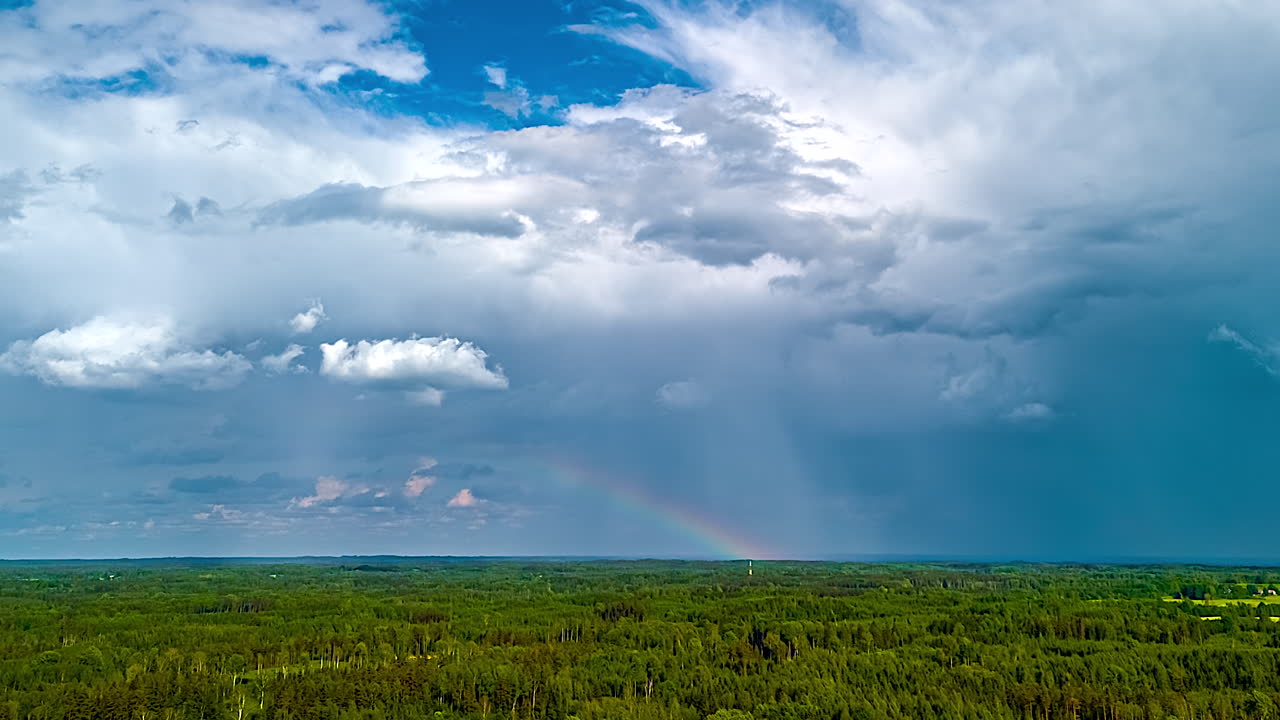 Rainbow appears in rain as clouds form and dissipate above remote green terrain in moody light and slow motion movement