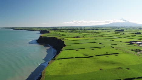 Vuelo-Aéreo-Sobre-El-Acantilado-Del-Océano-Con-Tierras-De-Cultivo-Y-Montañas-Detrás