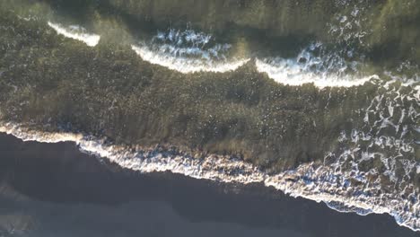 Vuelo-Con-Un-Dron-En-Ascenso-Con-La-Cámara-En-Lo-Alto-Viendo-La-Orilla-De-Una-Playa-Con-Las-Olas-Del-Mar-Alcanzando-Suavemente-La-Arena-Grisácea-En-Una-Tarde-De-Invierno-En-Valencia,-España