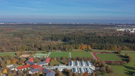 Wide-aerial-view-over-several-sports-grounds-with-the-skyline-of-Munich,-germany-in-the-background-behind-a-forest-landscape