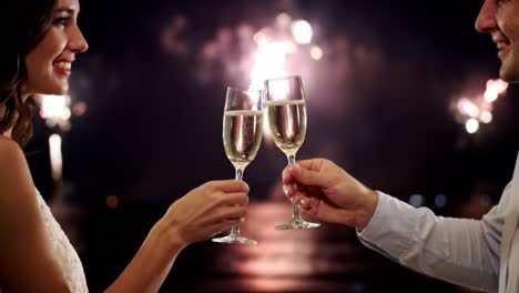 a couple celebrating a special moment together as fireworks illuminate the night sky, raising glasses of champagne in a romantic setting by the water