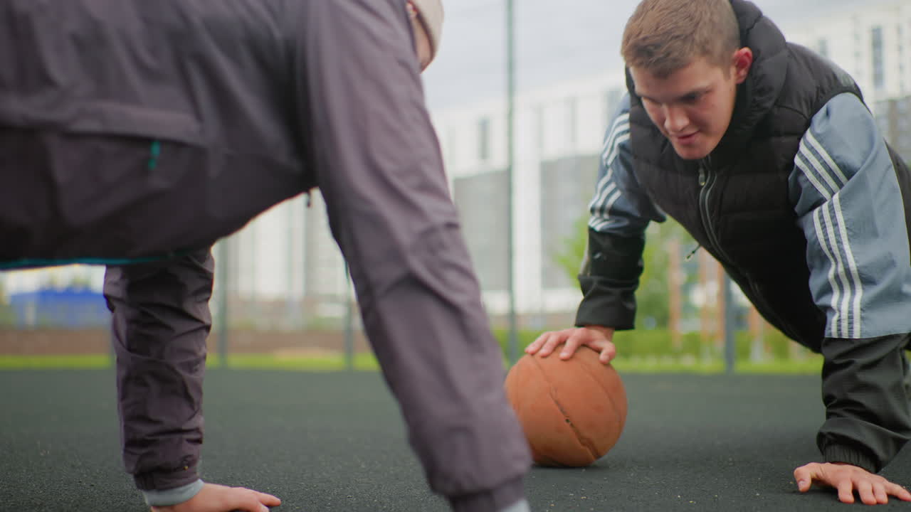 White man reaching basketball during pushup closeup on urban court, fierce expression and strained grip, competitor hand scrambling for ball, asphalt texture and hoop in background, mentorstyle
