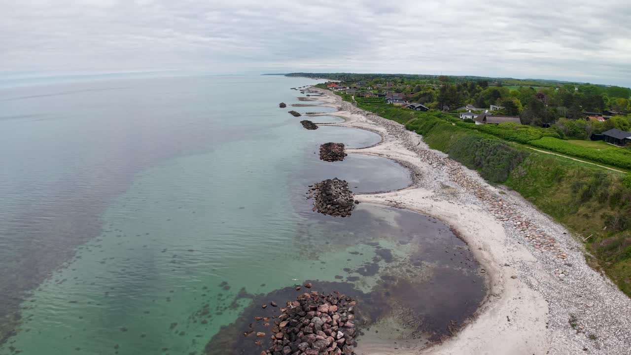 Flygvy med drönare över en stenig kustlinje med lugnt turkost vatten och en smal strand som möter gräsbevuxet land