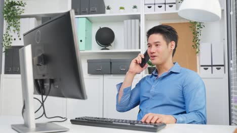 hombre de negocios asiático guapo hablando por teléfono móvil y trabajando en la oficina. joven empleado atractivo sentado en la mesa, escribiendo en la computadora portátil y discutiendo en el teléfono inteligente con colegas en el lugar de trabajo