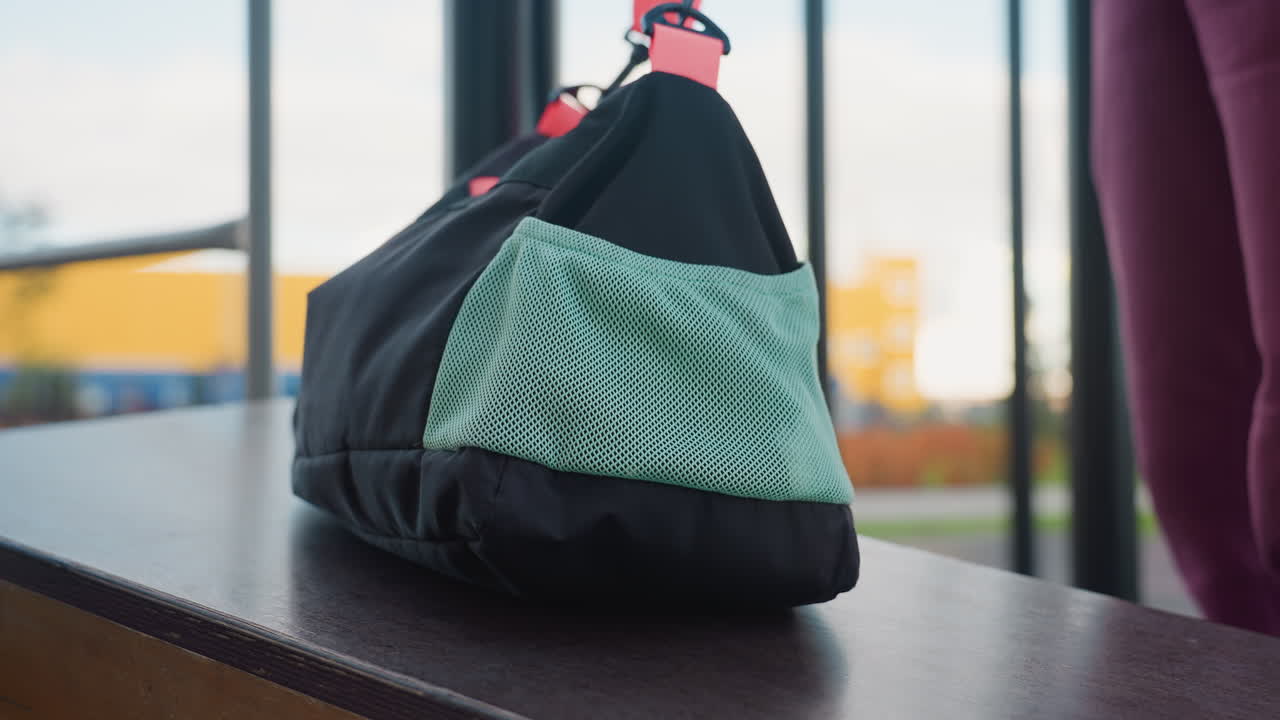 Youthful lady in magenta hoodie walks into urban gym area holding sports bag, places bag on corner of calisthenics bar, bends gracefully to tie shoelace on bar before starting workout under open sky