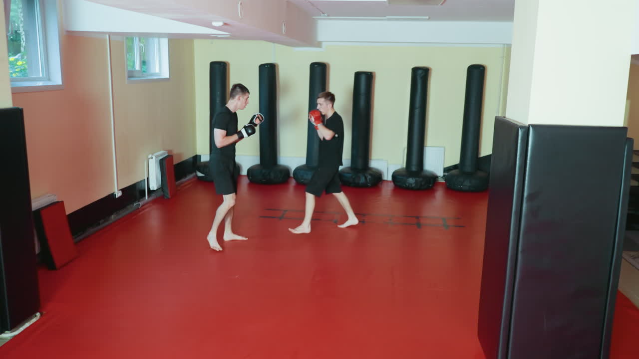 레슬링 선수들이 무술 체육관의 빨간 매트 위에서 맨발로 스파링을 하고 있어요. 한 명은 빨간 글러브로 얼굴을 가리고 있고, 다른 한 명은 공격을 준비하고 있어요. 훈련장 안에는 서 있는 샌드백들이 있고, 격투 기술을 연습하고 있어요.