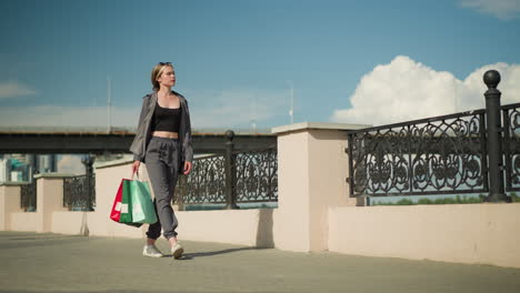 lady in casual grey outfit walking near a bridge fence, carrying three colorful shopping bags in hand, cars are visible passing on the overhead bridge in the background, with a clear blue sky