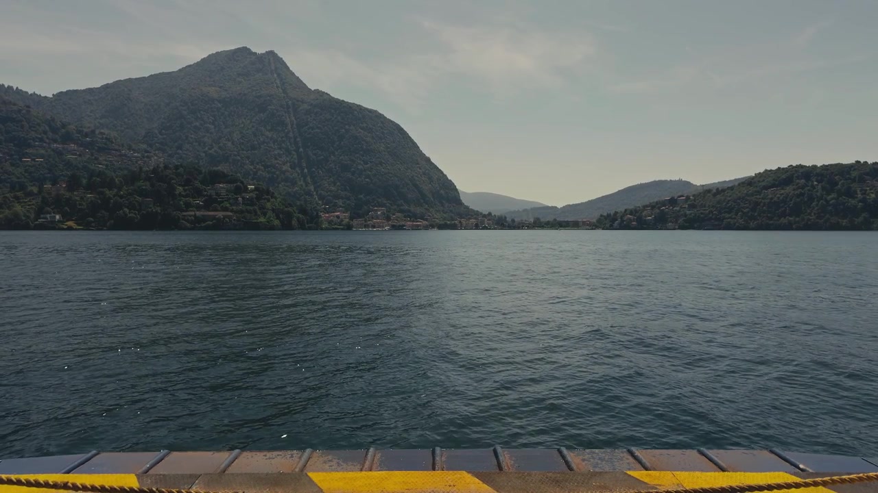 Vista dalla rampa di carico di un traghetto in movimento attraverso un lago calmo con acqua blu profonda e montagne alpine mozzafiato, che cattura un viaggio panoramico in traghetto attraverso il nord Italia.