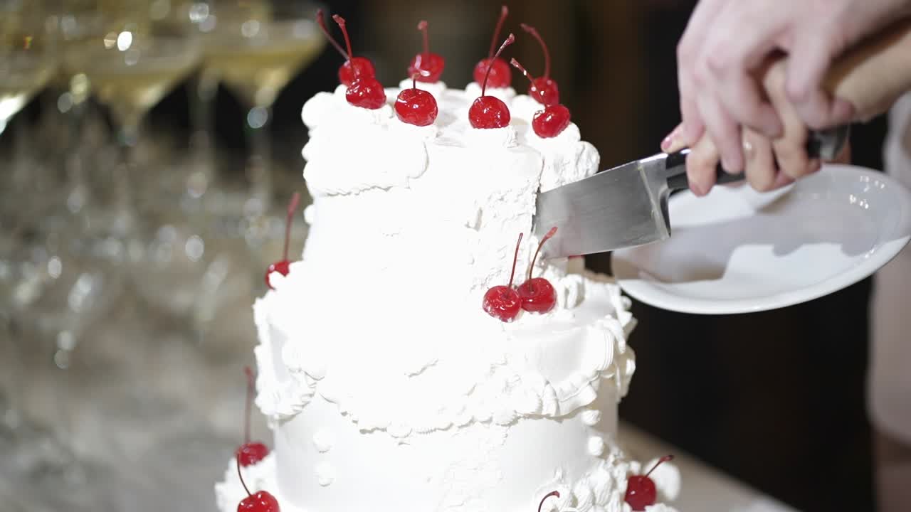 Close-up of hands cutting an elegant white wedding cake topped with cherries, illuminated by a bright flash from the photographer during the reception, slow motion shot