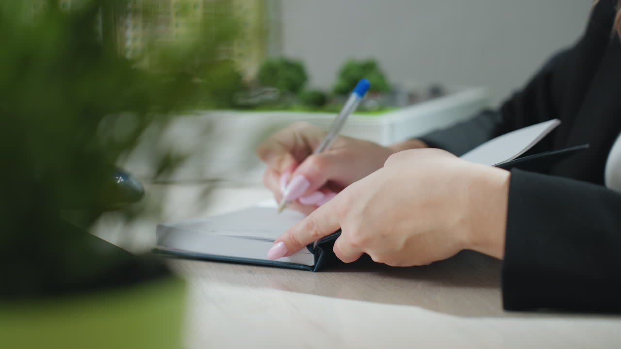 close up partial view of fair skin lady with polished pink nails writing in journal using pen on wooden desk with blurred office background