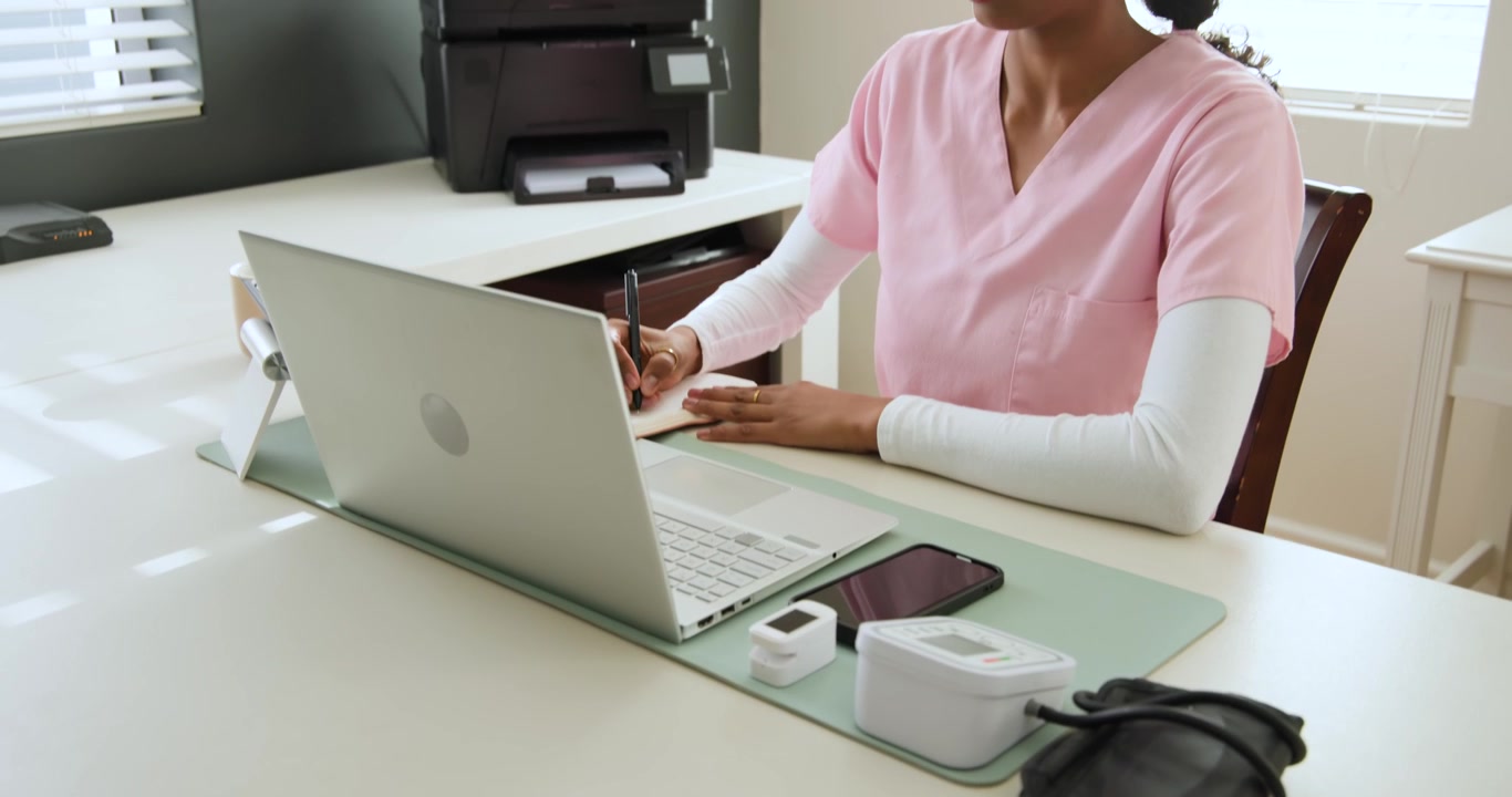 Donna afroamericana in camice rosa alla scrivania, con il laptop aperto, che prende appunti, digitando per una visita virtuale.