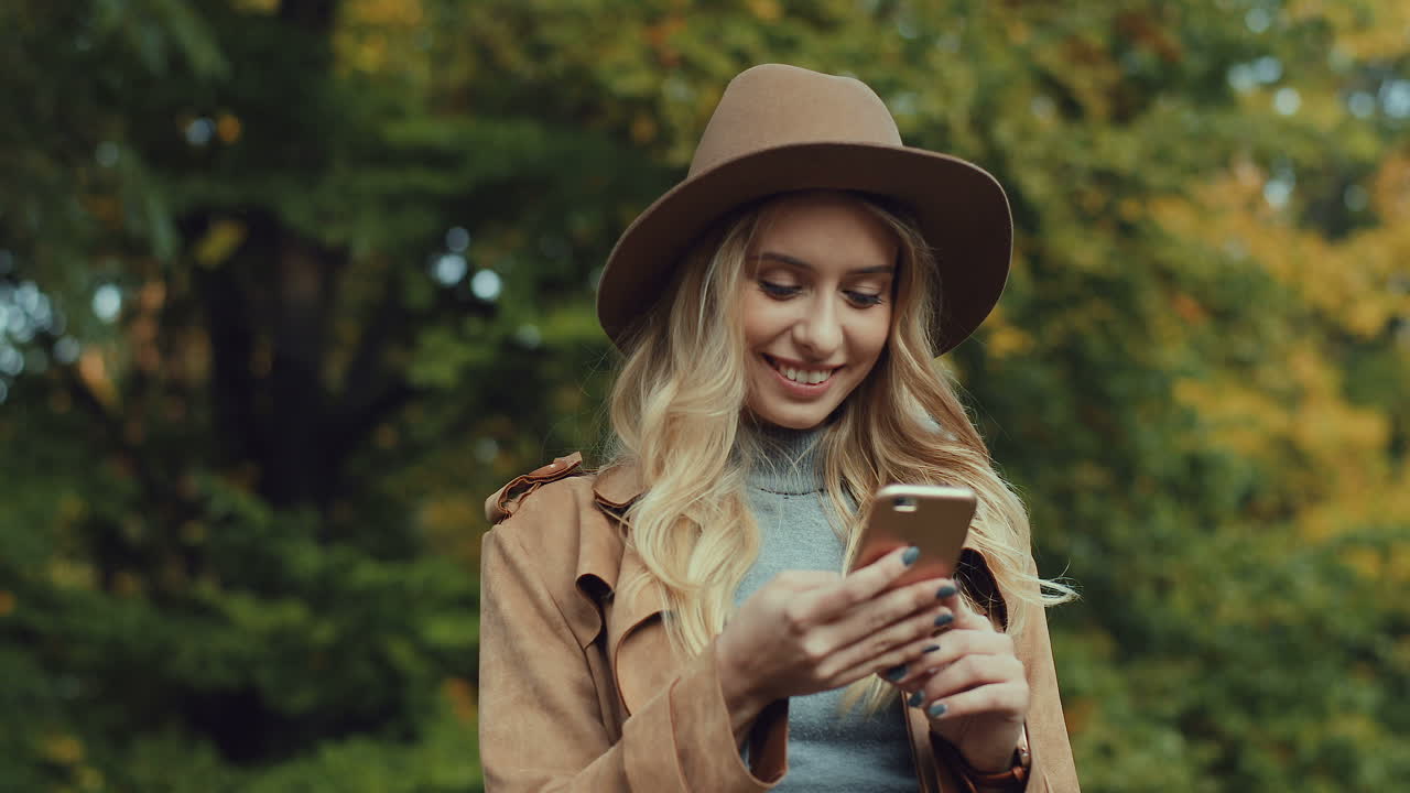 Caucasian young blonde woman wearing a hat and texting on smartphone in the park in autumn