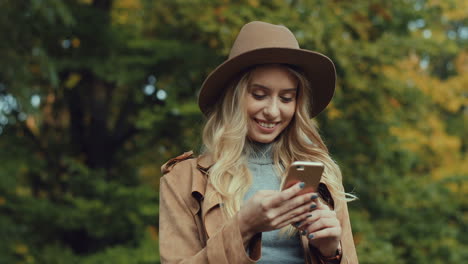Caucasian-young-blonde-woman-wearing-a-hat-and-texting-on-smartphone-in-the-park-in-autumn
