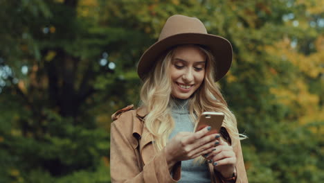 caucasian young blonde woman wearing a hat and texting on smartphone in the park in autumn