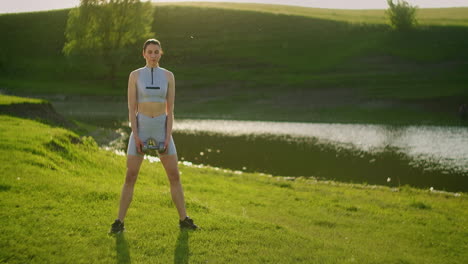 a woman squats with dumbbells at sunset in a park against the backdrop of a river. outdoor training.