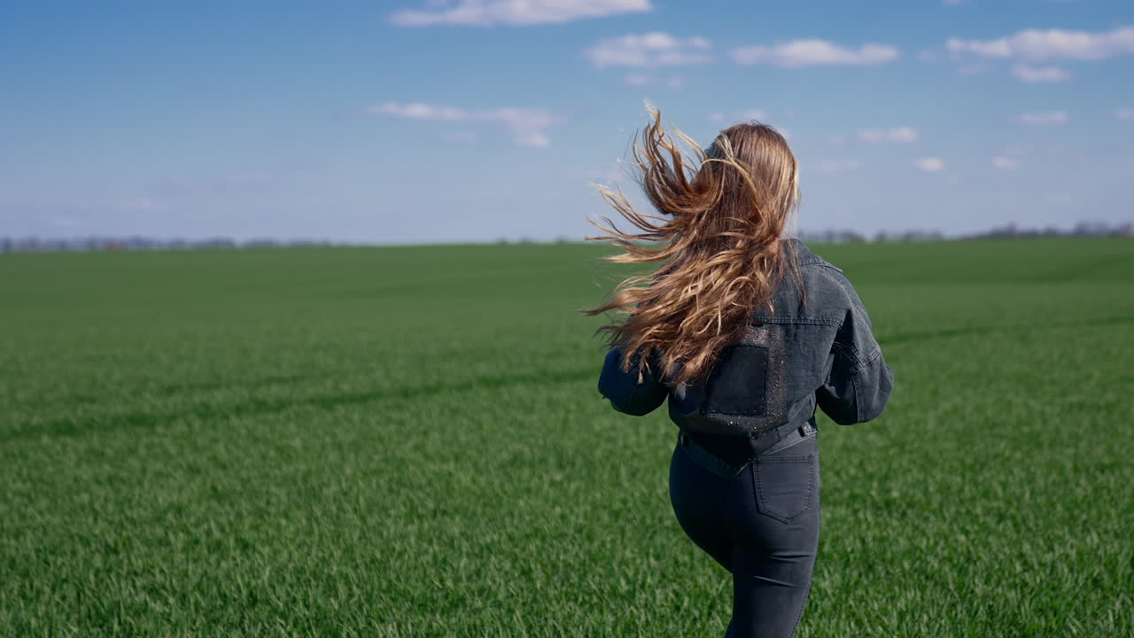 Girl is running on green field. Young woman with long fluttering hair in denim suit runs in nature and turning her cheerful face on camera. Rear view. Slow motion.