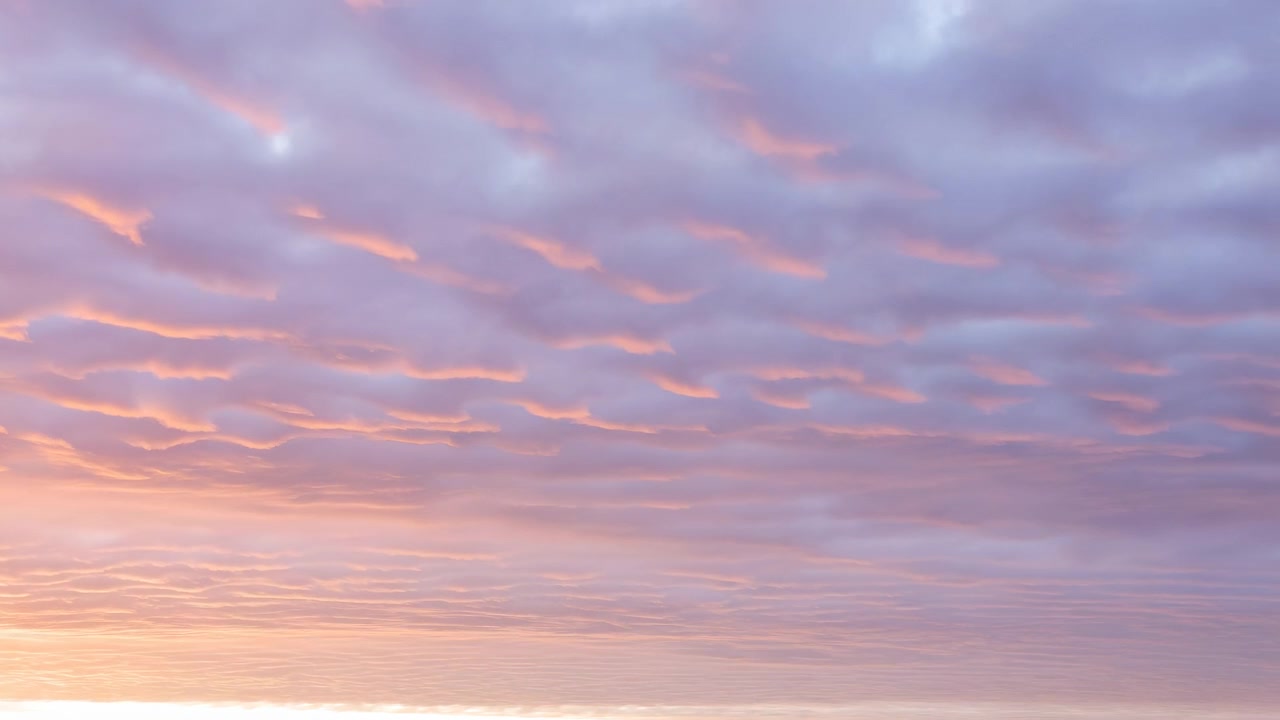 Expansive stratiform cloud deck fills wide sky, lit from below by warming sunrise glow that lifts peach and gold highlights along undersides while upper layers stay soft lavender and calm