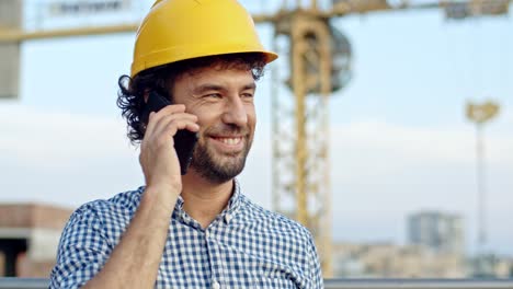 primer plano del joven caucásico apuesto en el casco amarillo hablando por teléfono y sonriendo en el sitio de construcción. al aire libre.